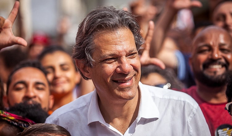 Haddad em SJC participará de ato de recepção e sabatina, na foto ele olha para a direita, com uma camisa branca, sorrindo, com pessoas ao fundo, fazendo um L com as mãos.