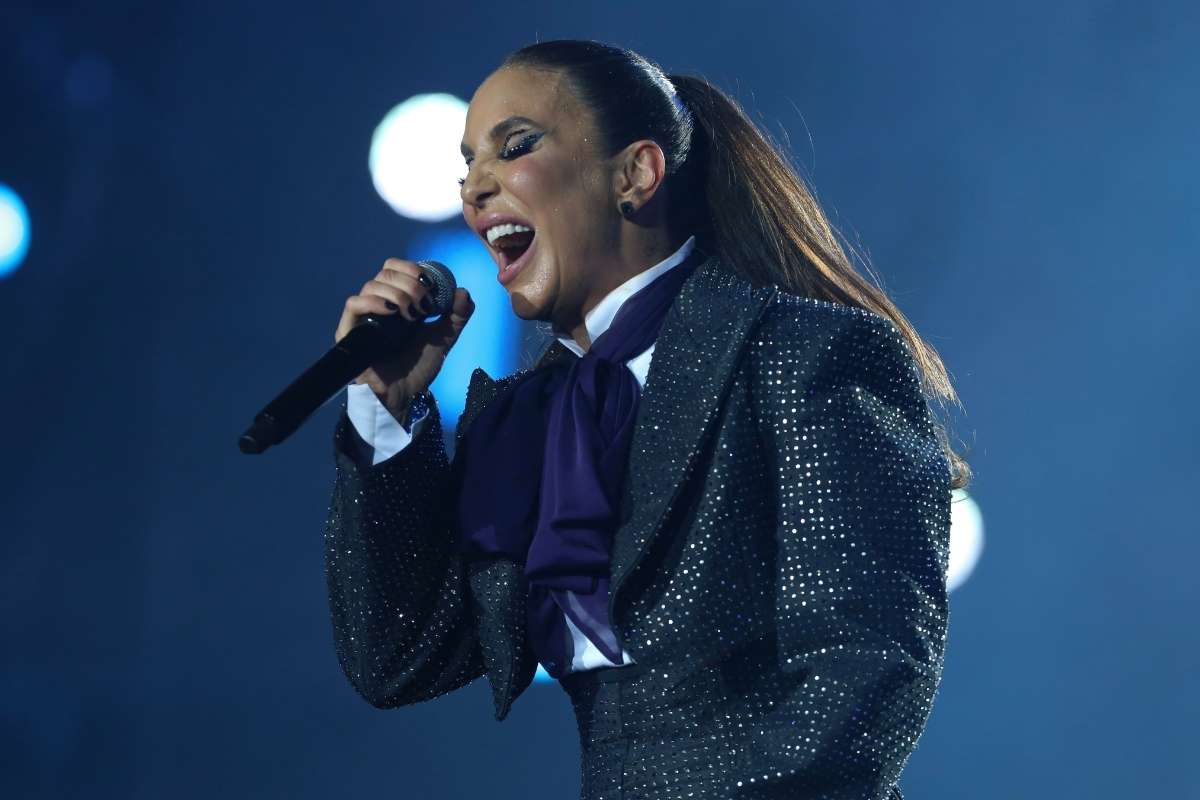 Ivete Sangalo com um microfone na mão, durante o Rock in Rio, com um fundo azul e uma roupa preta.