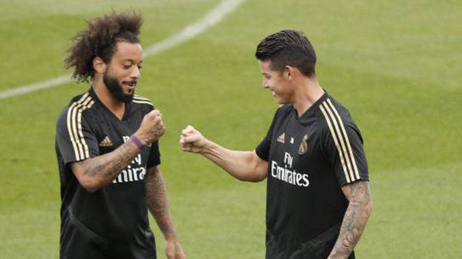 Jogador Marcelo ao lado de James Rodríguez, com a camisa preta do Real Madrid. Ambos fazem um sinal com as mãos; ao fundo o gramado verde.