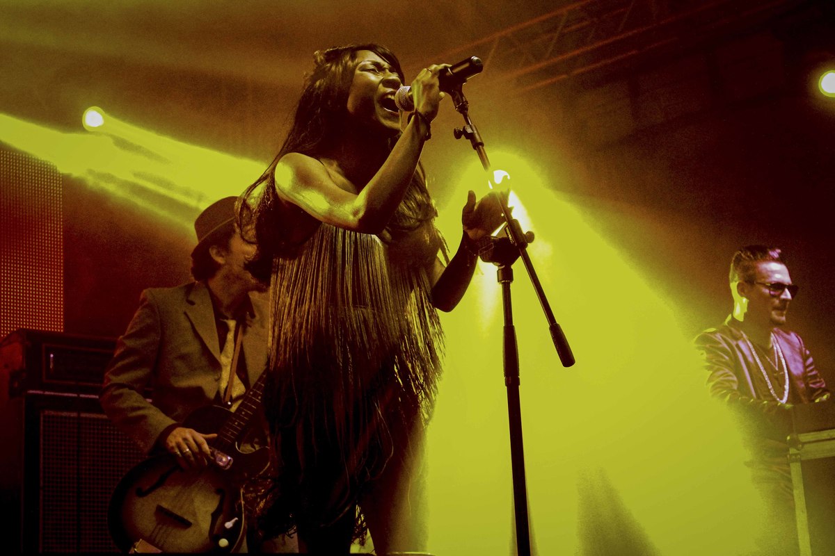 Cantora moçambicana Koko Jean Davis em apresnetação. Ela canta segurando um microfone em pedestal e tem expressão potente e enérgica.