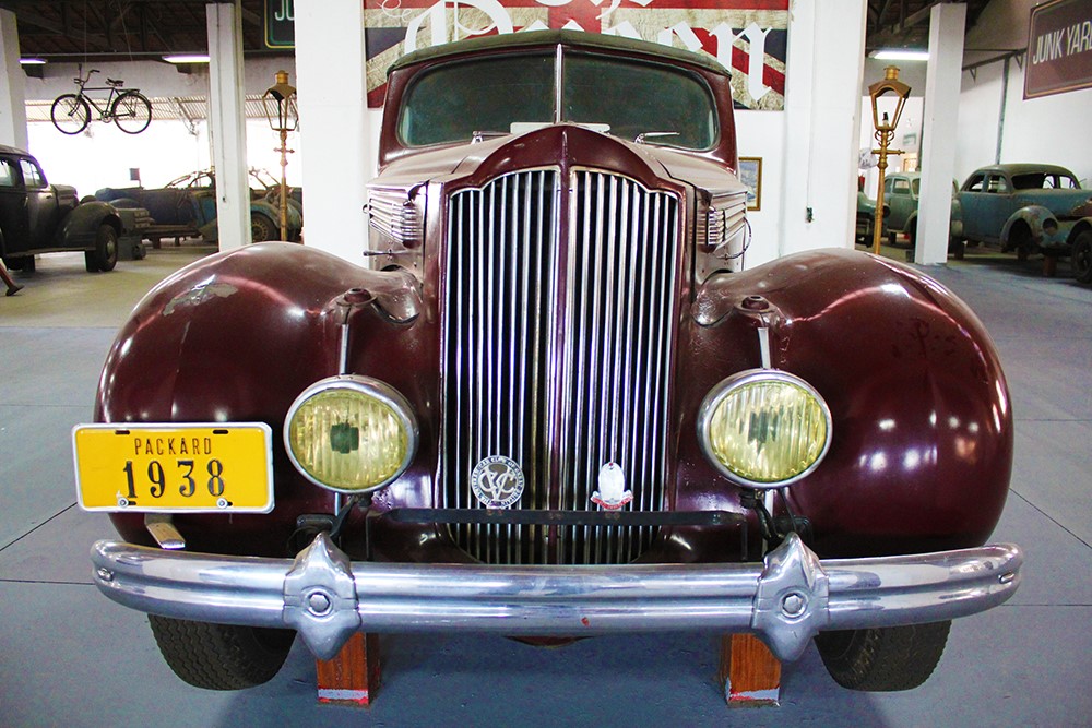 Carro bordô, modelo Packard 1938, exposto no Museu Roberto Lee, de frente para a câmera, com uma placa amarela e o museu ao fundo, com menos detaque.
