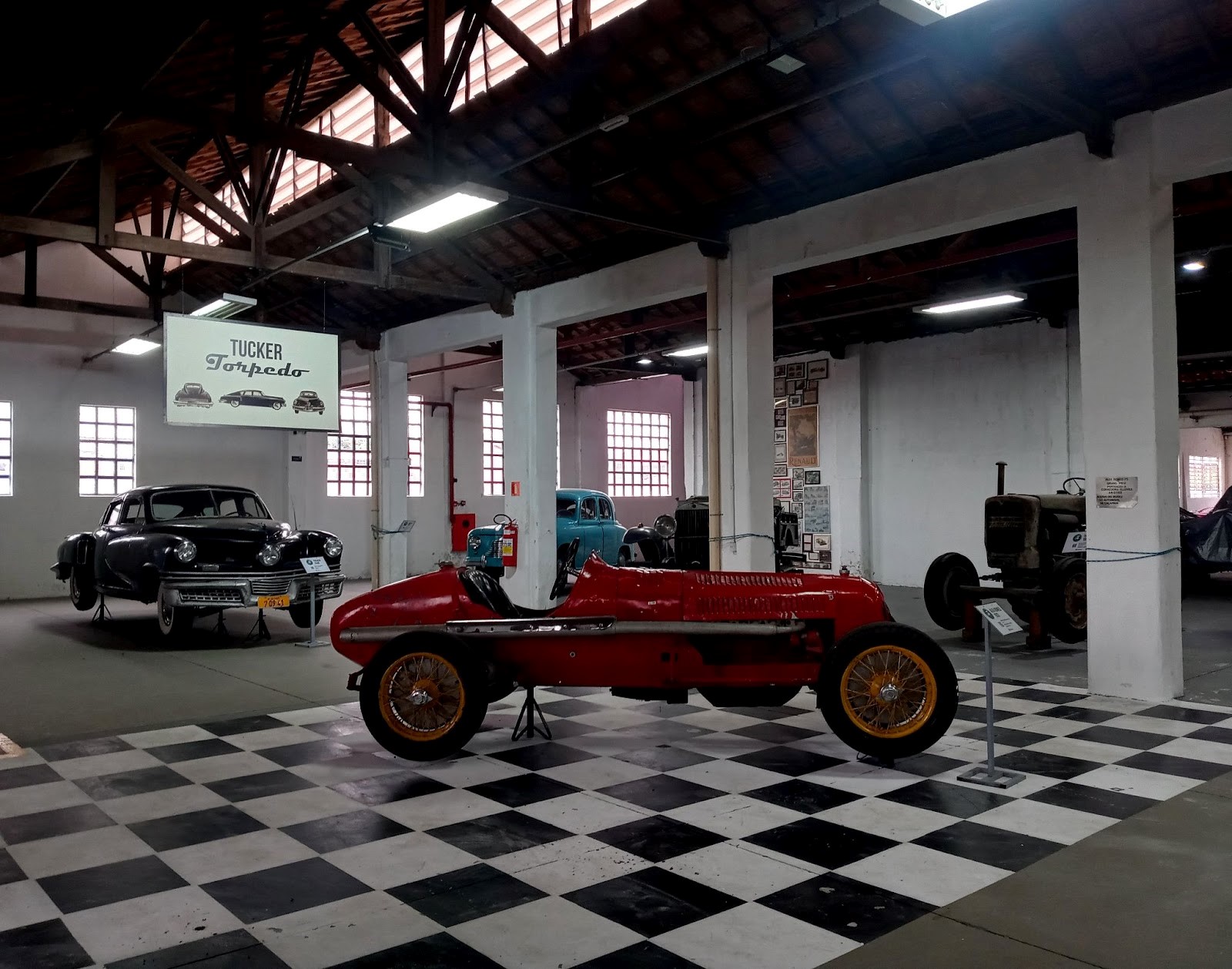 Museu Roberto Lee, um dos pricipais pontos turísticos de Caçapava, com um carro vermelho no centro, um preto à esquerda, um azul ao fundo, pretos à direita, chão quadriculado preto e branco, um teto de madeira e uma placa ao fundo como nome do local.