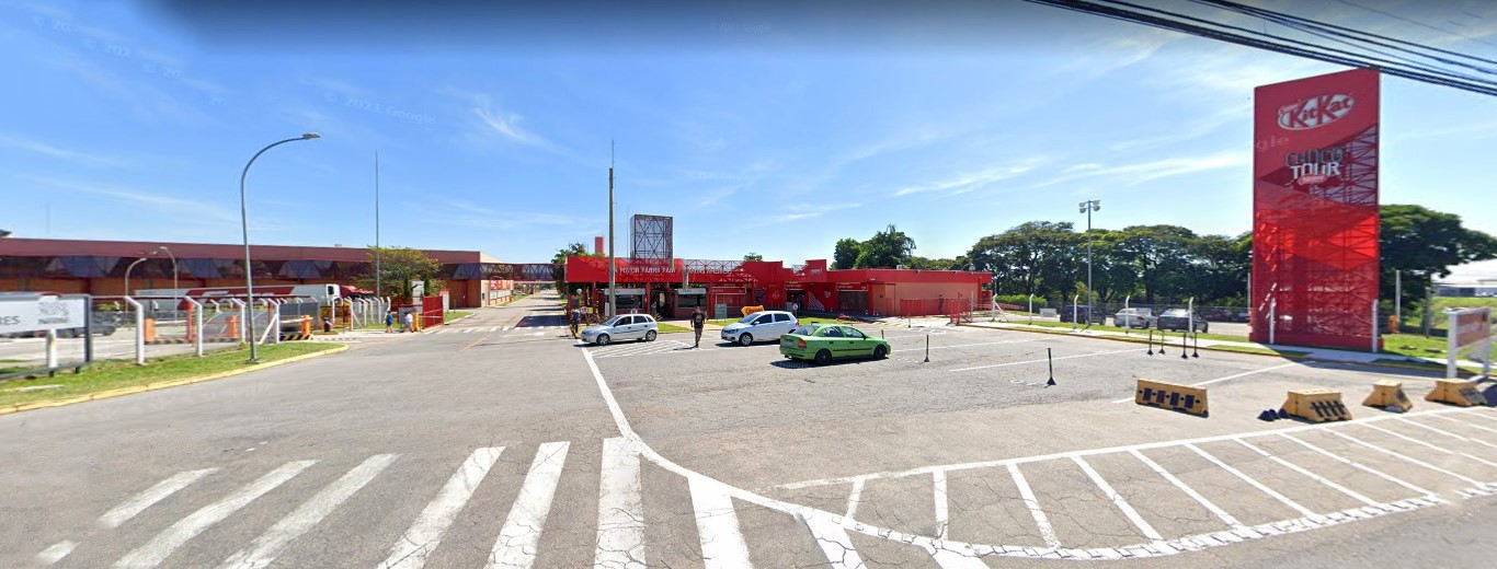 Entrada da fábrica Nestlé, em Caçapava, com destaque para a larga rua e o prédio em vermelho, com placas grandes com o nome, além de carros estacionados em frente.