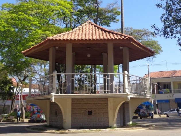 Coreto da Praça da Bandeira, um dos pontos turísticos de Caçapava, no meio de uma praça, com telhado marrom, pilastras brancas, grades brancas e casas em volta.