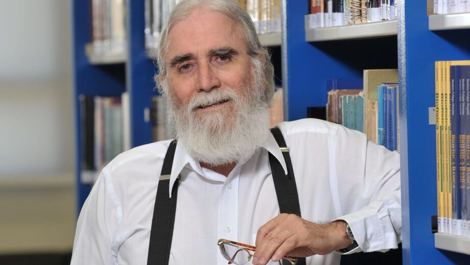 Professor Justino, referência em eletromagnetismo, será homenageado pelo Parque Tecnológico de São José dos Campos (SJC); na foto ele usa uma camisa branca, um suspensório preto e sorri para a câmera, na frente de livros.