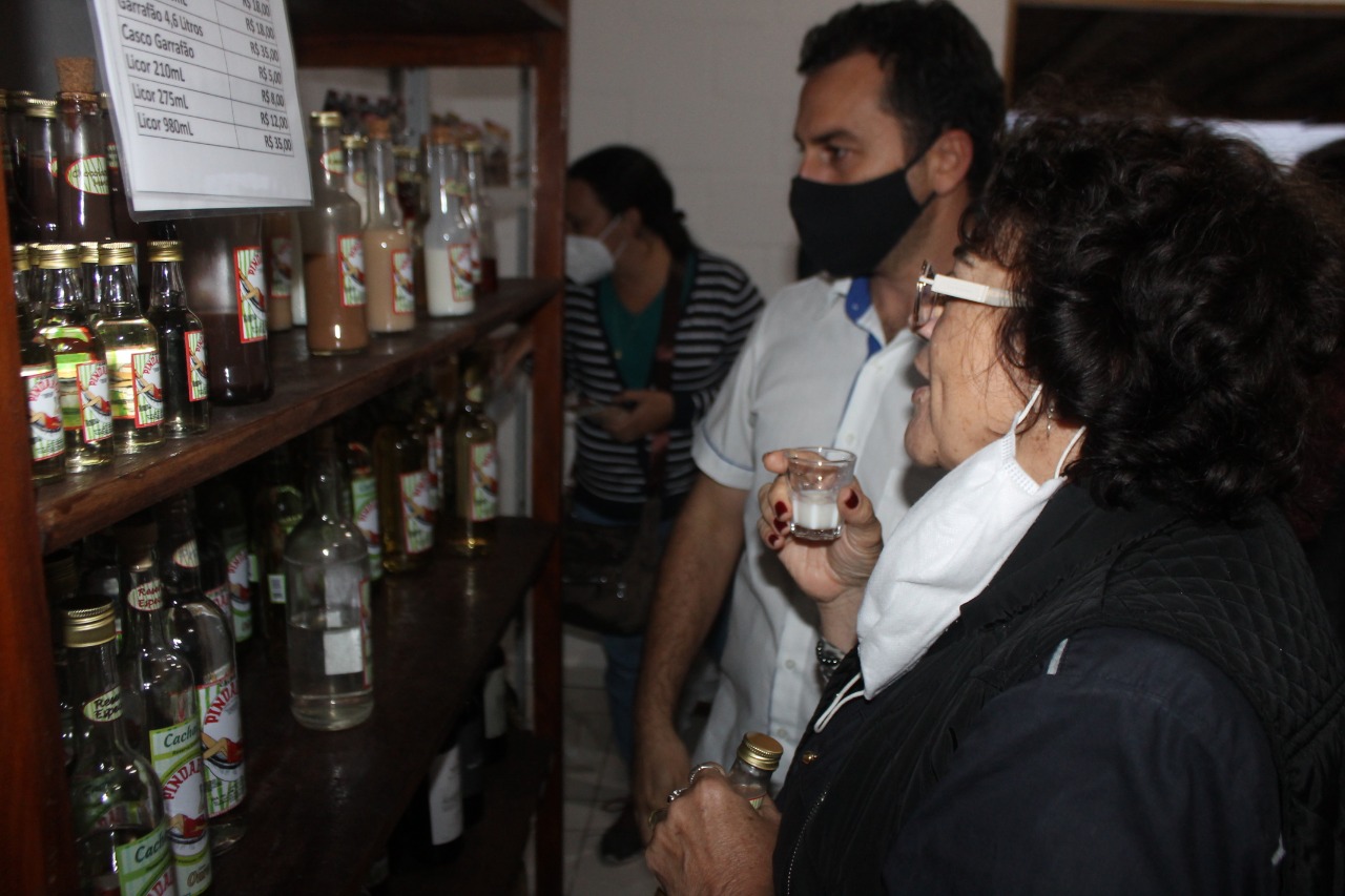 Mulher provando uma cachaça durante a Semana do Turismo de Pindamonhangaba, em frente a uma estante com garrafas.