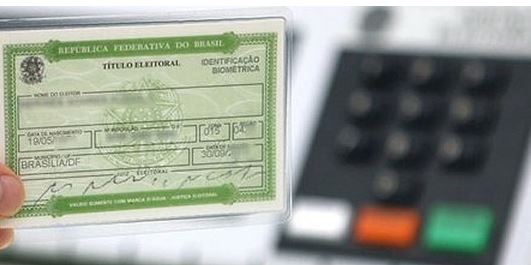 Título de eleitor em destaque, na frente da foto, na cor verde; ao fundo a urna eletrônica, com os botões coloridos, desfocada.