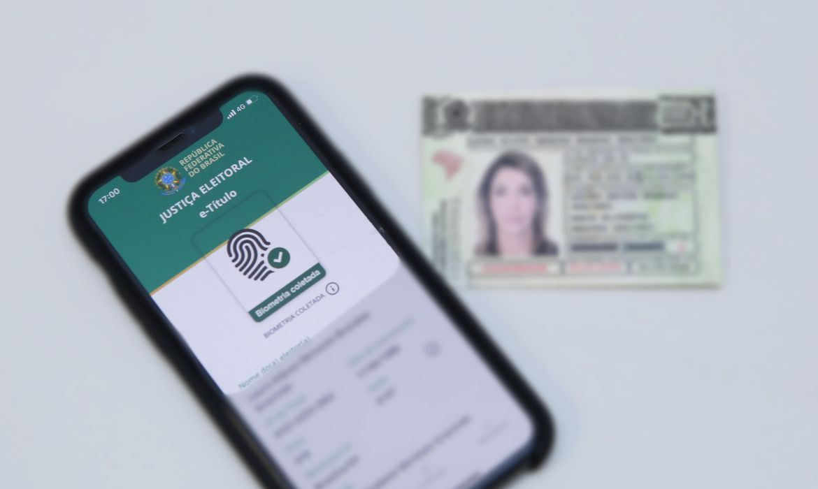 Imagem mostra celular com E-título aberto e título de eleitor físico ao lado.