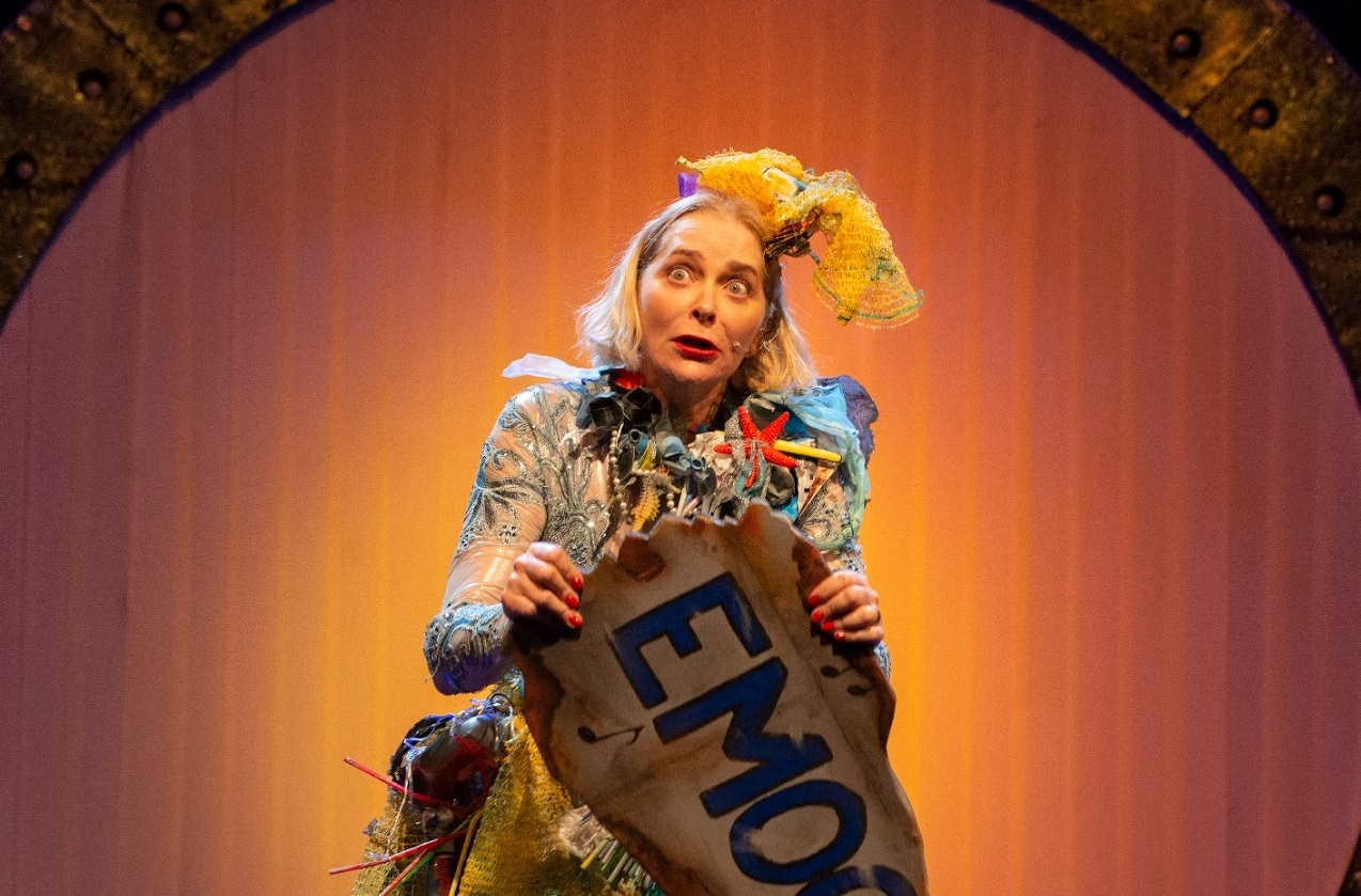 Na Foto, Alexandra Richter, que se apresenta no Teatro Colinas com comédia Uma Loira na Lua ; foto mostra fundo laranja e a atriz com um traje próprio do espetáculo, segurando uma placa.