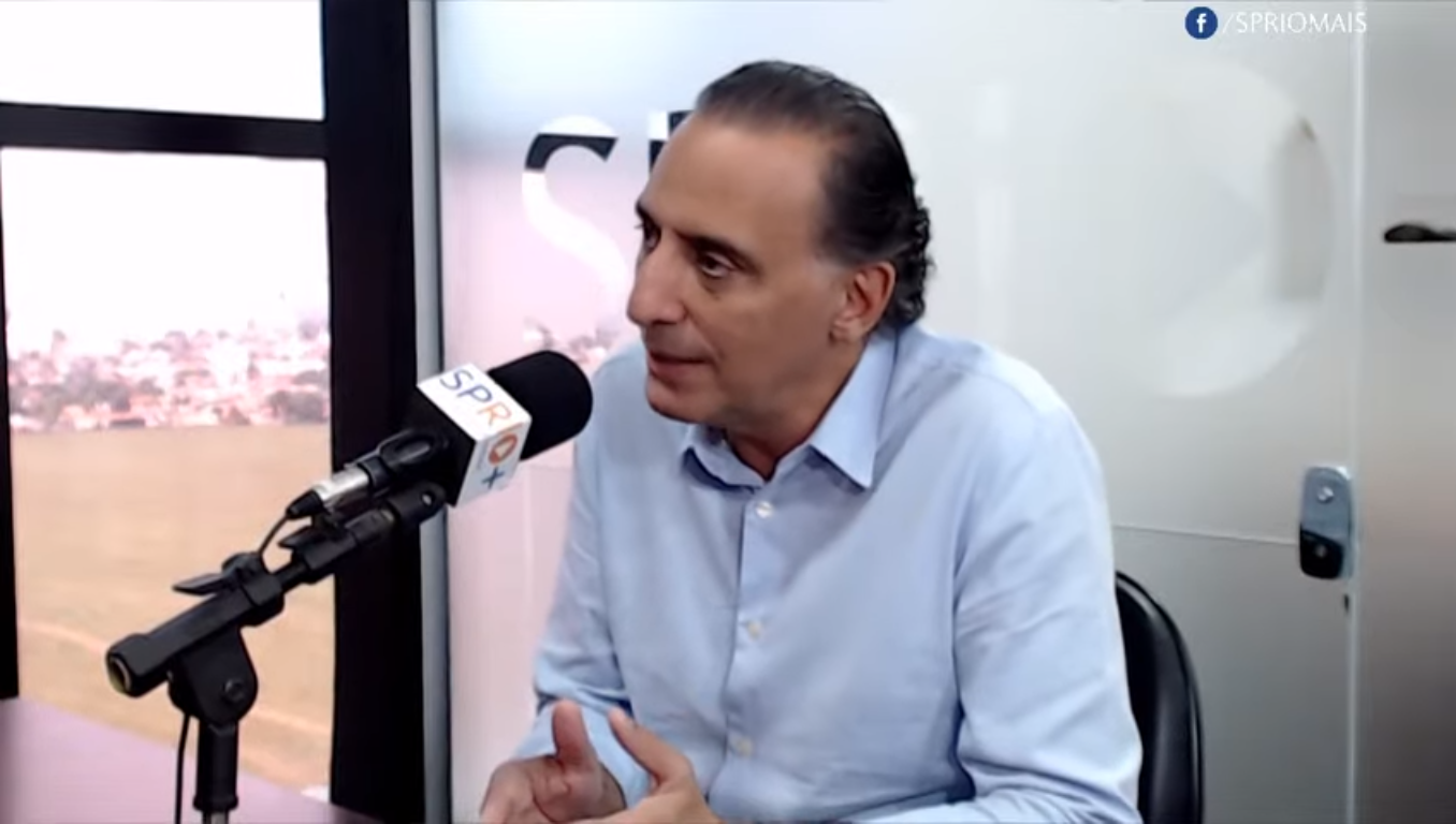 Eduardo Cury durante entrevista para a SP RIO+, com um microfone, sentado na cadeira, vidro ao fundo a vista de São José dos Campos.