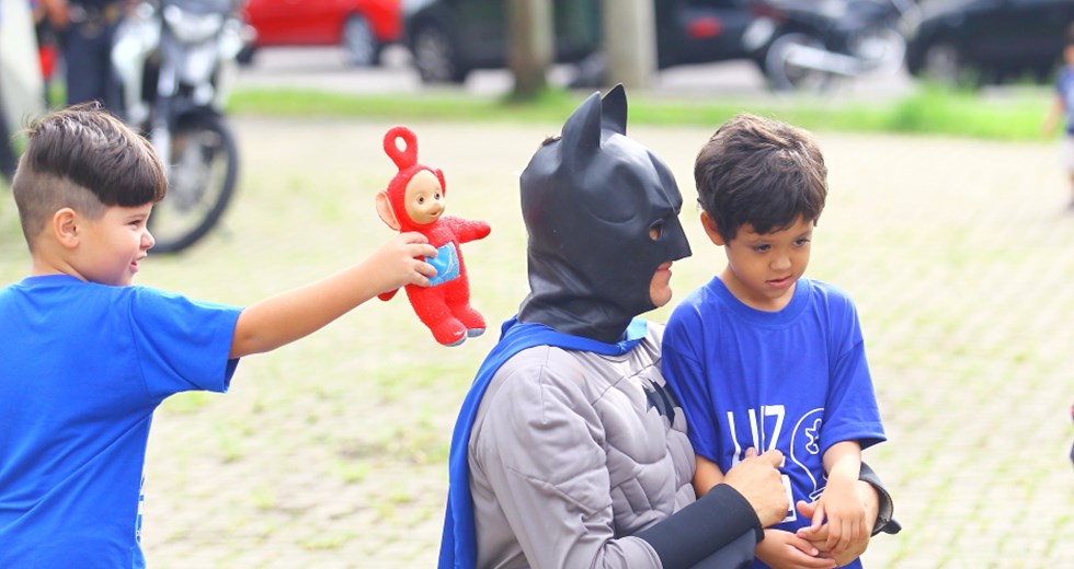 Crianças com camiseta azul, uma delas segura um boneco vermelho, próximo a um homem com fantasia de Batman, durante o evento Blue Day, voltado para crianças autistas.