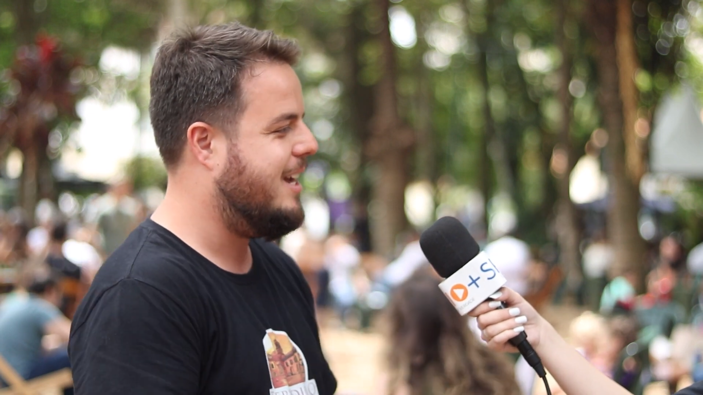João Guilherme Mascarenhas, proprietário da Torradillo Café, durante entrevista à SP RIO+, falando no microfone, com área verde ao fundo e camiseta preta., durante o Destination Gastronomia.