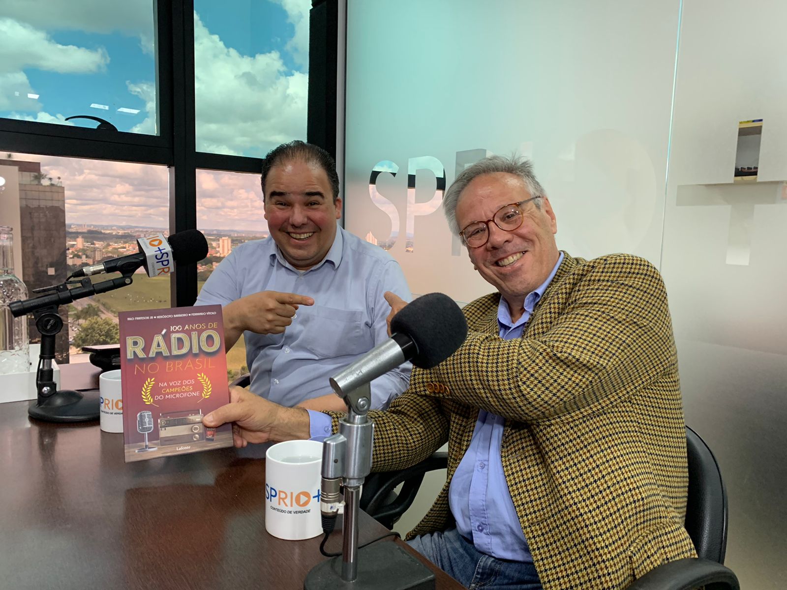 Foto mostra Claudio Nicolini, jornalista da Band Vale, à esquerda, sentado, e Nilo Frateschi, segurando seu livro 100 Anos de Rádio no Brasil Na voz dos Campeões do Microfone, à direita; ao fundo a cidade de São José dos Campos; no meio uma mesa com o microfone da SP RIO+, do podcast Talk+.