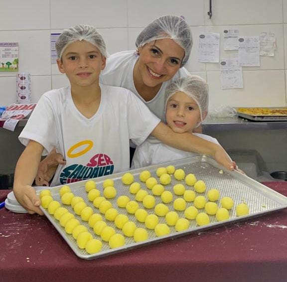 A sócia proprietária da Santo Sabor Saudável, Eliana Duarte, no meio, ao lado de seus netinhos, com uma bandeja de brigadeiros, camiseta branca e touca, em uma cozinha. / Foto: Divulgação