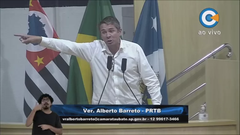 Vereador Alberto Barreto (PRTB), de Taubaté, falando sobre rodeio na cidade, com camisa branca, apontando para o lado e bandeiras no fundo.