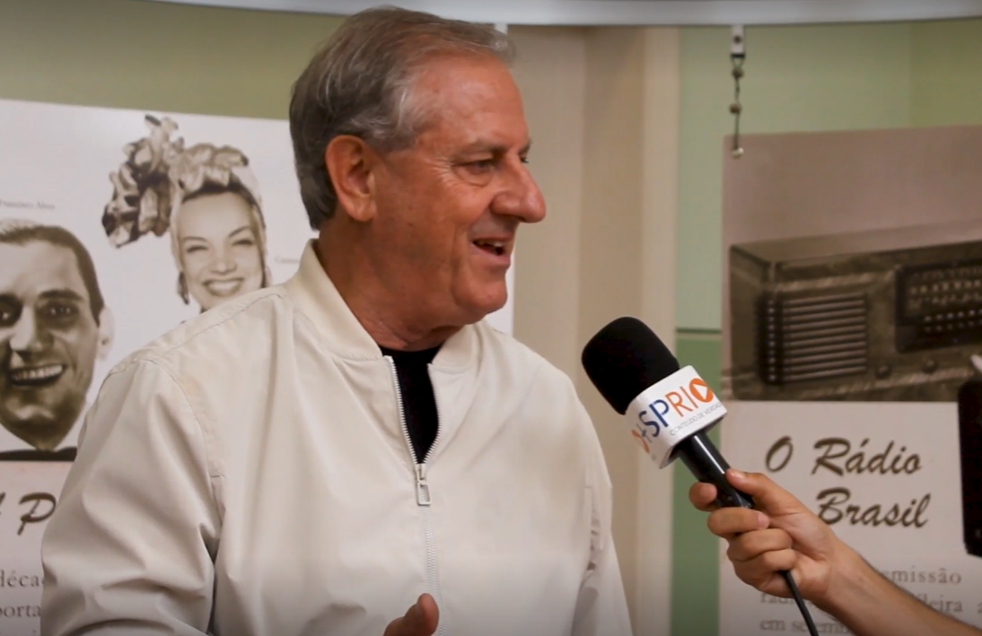 Aldo Zonzini, presidente da AFAC, em entrevista à SP RIO+, com uma roupa branca, olhando para a direita, falando no microfone, sorrindo.