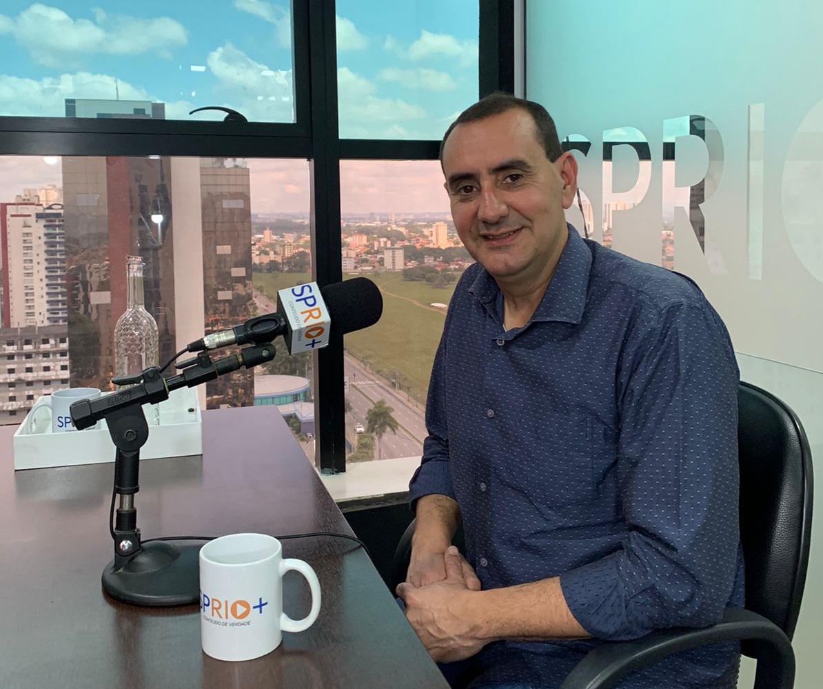 Retrato do Anderson Farias, do PSD, prefeito de São José dos Campos, durante entrevista à SP RIO+, sentado numa mesa, com uma caneca, um microfone, a paisagem ao fundo, parede vidro e uma janela, falando sobre a municipalização das escolas estaduais da cidade.