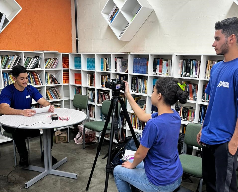 Associação Celebreiros realizando workshop de produção audivisual, com camisetas azul, parede branca e laranja, prateleiras e estantes com livros.