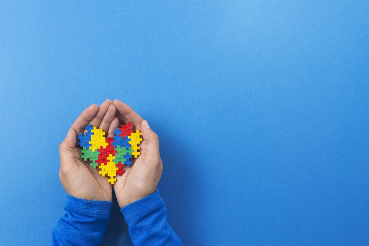 Mão segurando um coração de quebra cabeças coloridos, símbolo do autismo, com um fundo azul.