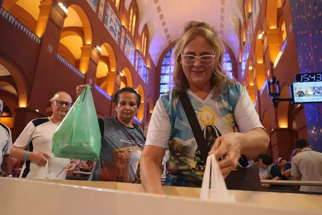 Mulher com a camiseta de Nossa Senhora Aparecida, segurando uma sacola, fruto de uma doação, com a Basílica ao fundo e outros fieis ao lado.