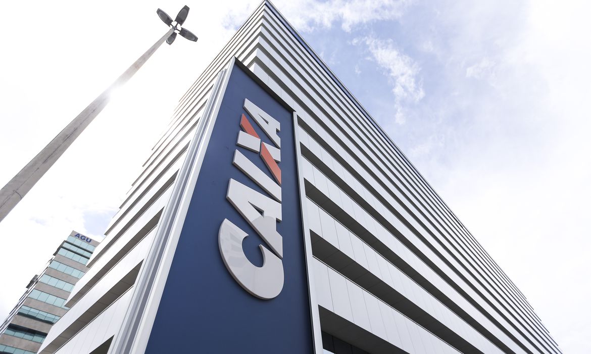 Fachada da Caixa Econômica Federal, pagadora do abono salarial, com céu azul e poste ao lado.