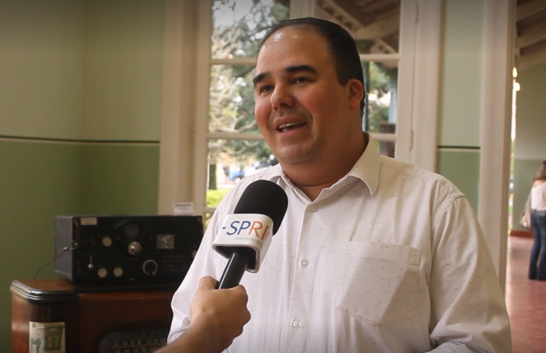 Claudio Nicolini durante entrevista ao portal SP RIO+, com camisa branca, sorrindo, falando no microfone, durante exposição dos 100 anos do rádio, no Parque Vicentina Aranha, com o cenário do parque ao fundo.