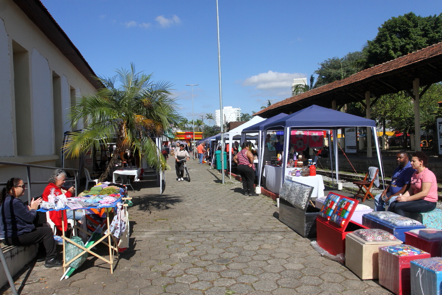 Barracas na Feira dos Trilhos, em Jacareí, em uma rua, com o céu azul ao fundo, pessoas andando.