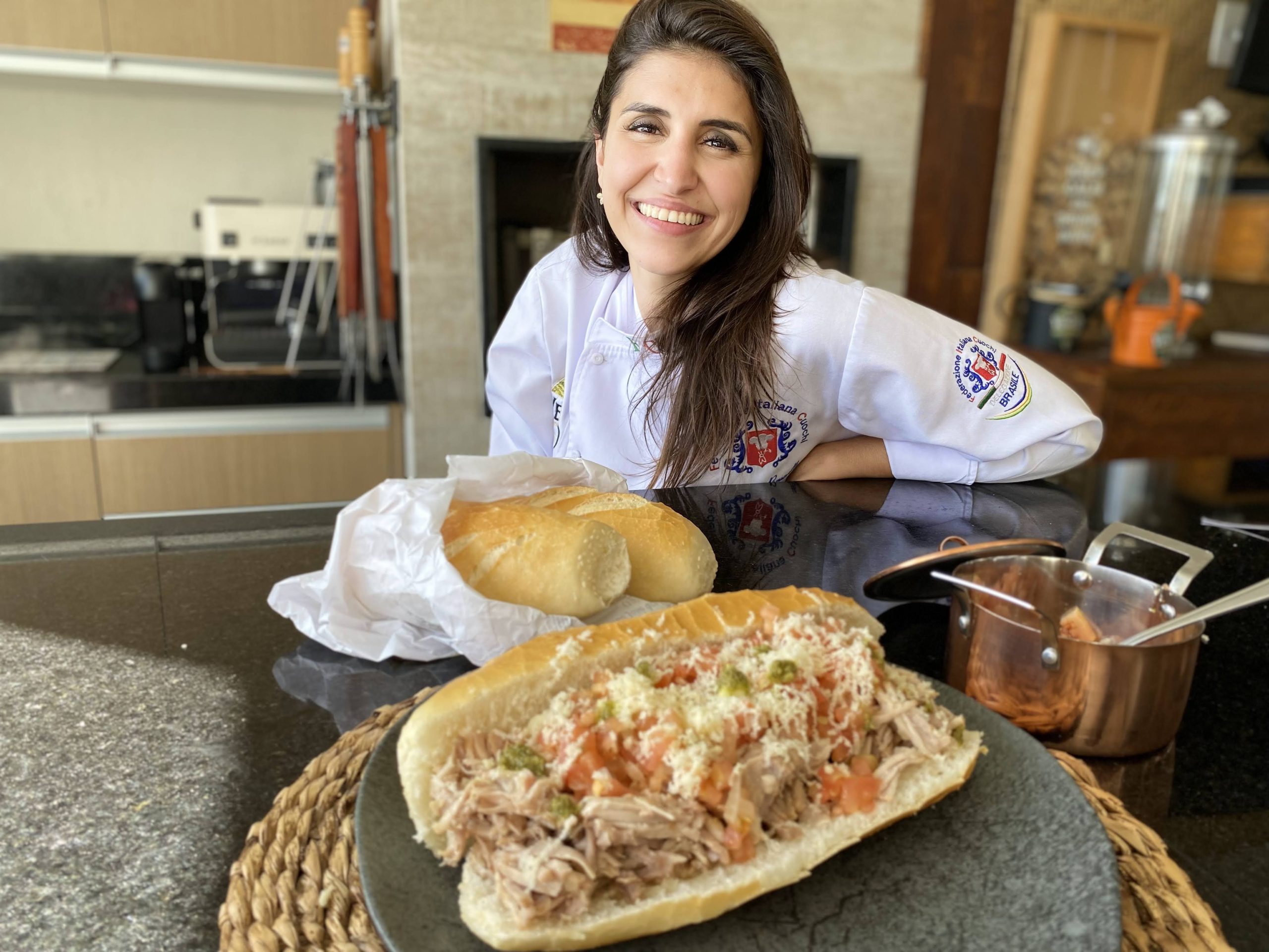 Chef Flávia Brunelli, fundadora da Del Veneto, olhando para a câmera, sorrindo, com roupa branca, em frente a uma mesa com comidas em cima, e uma cozinha ao fundo. Marca estará no Destination Gastronomia, em São José dos Campos.
