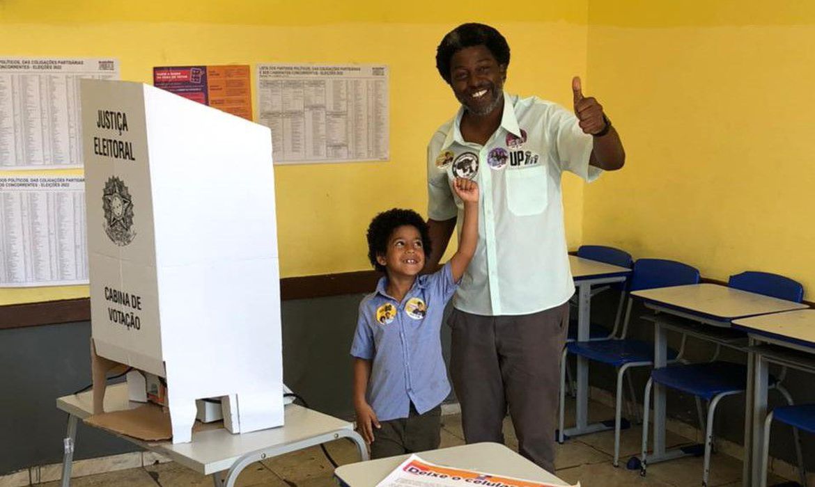 Leonardo Pericles votando, com uma camisa clara, calça cinza, ao fundo de uma parede amarela e com uma criança ao lado.
