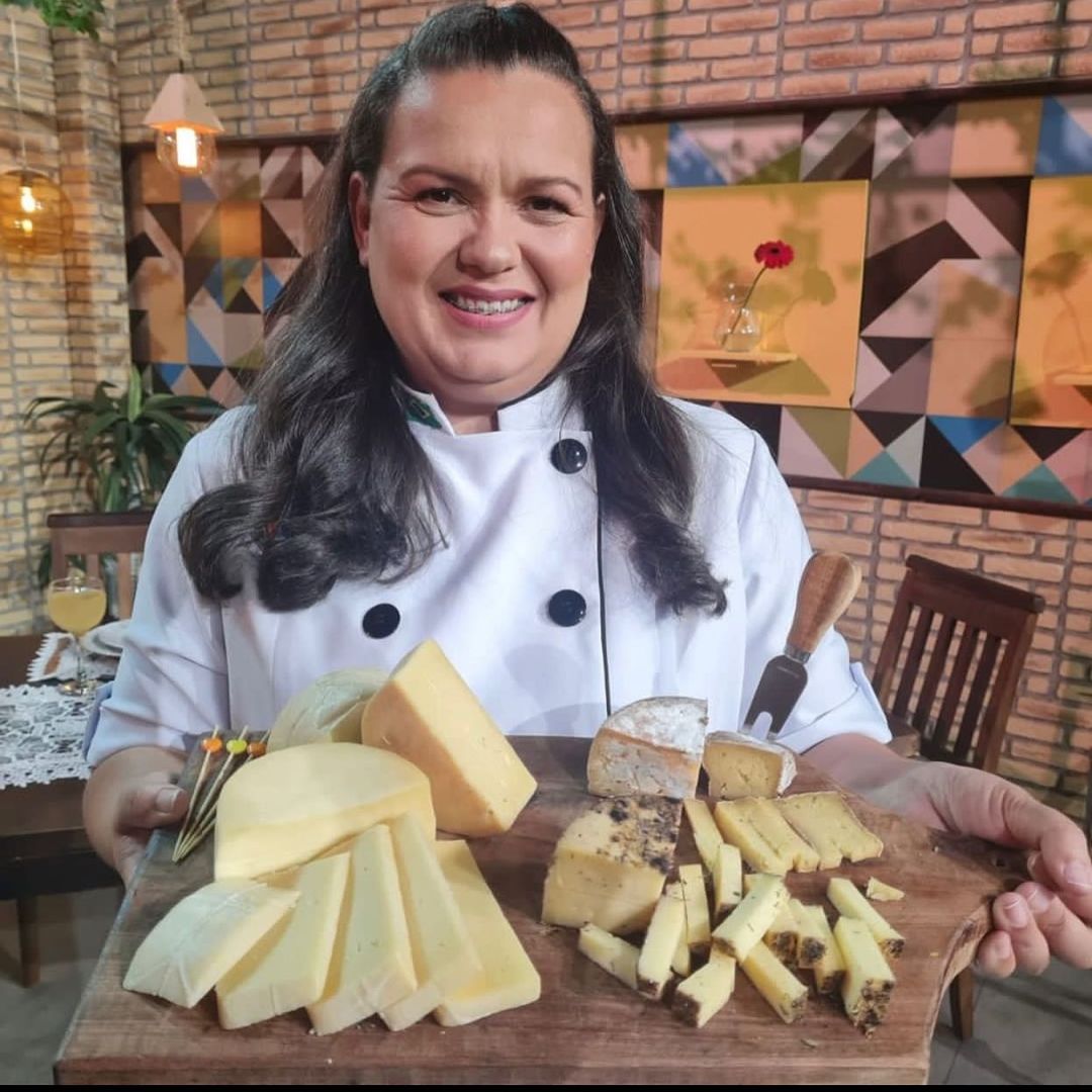 Foto mostra Camila Almeida, queijeira de Caçapava, segurando uma tábua com diversos queijos, com um jaleco branco e um fundo marrom, ilustrando sua participação na 2ª Edição do Mundial do Queijo Brasil.