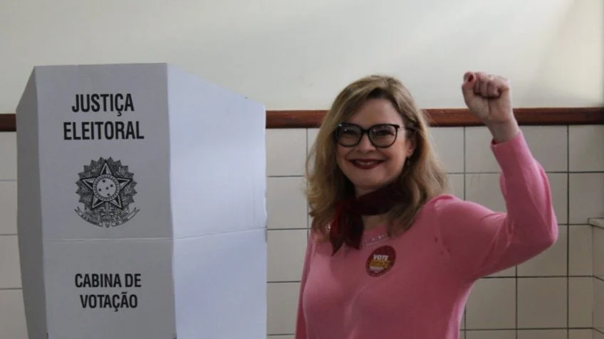 Sofia Manzano votando, ao lado da urna, com uma camisa rosa e com a mão para cima.