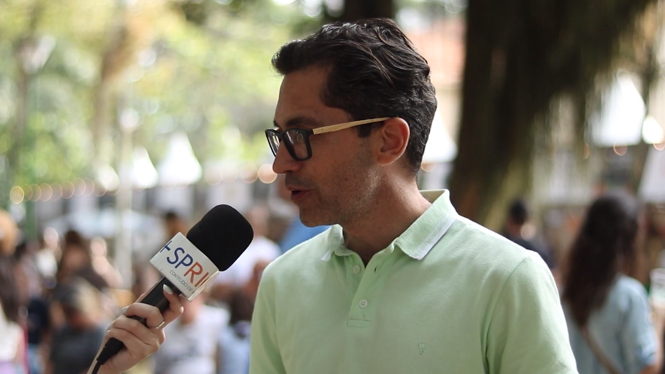 Tom Freitas, presidente da FCCR, durante entrevista à SP RIO+, com microfone, camisa verde, fundo de natureza, durante o Destination Gastronomia.