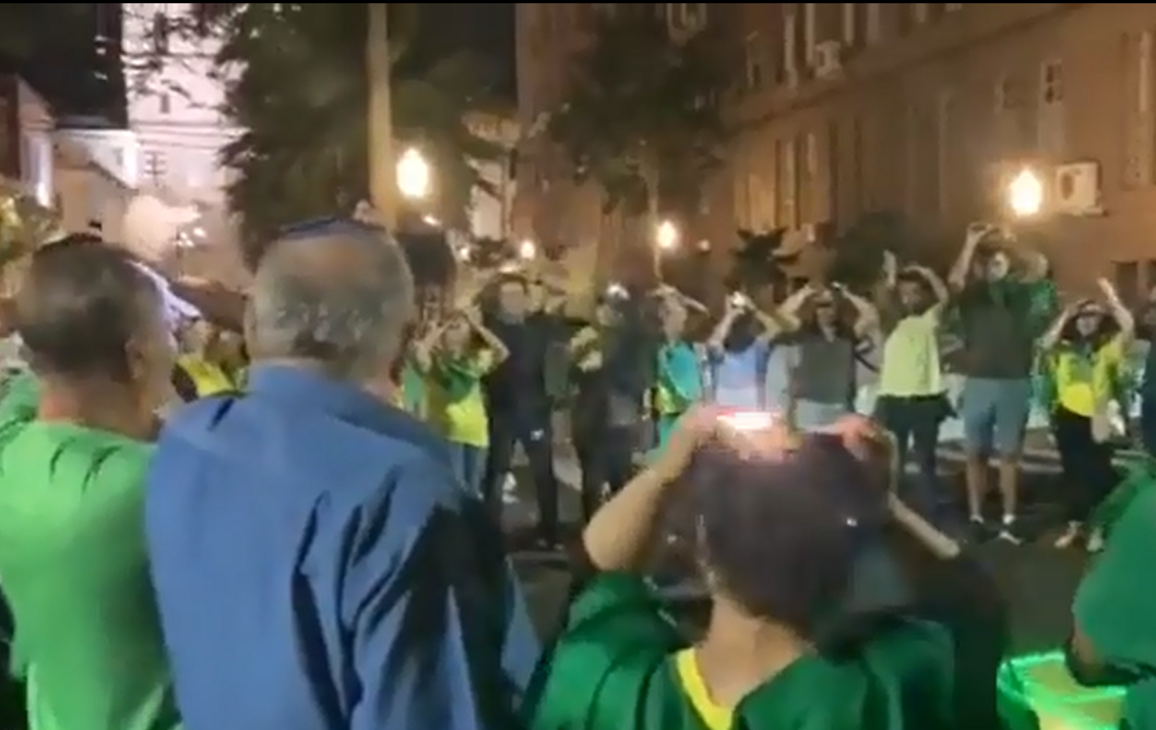 Multidão de bolsonaristas pedindo ajuda a 'extratrerrestres', com celular na cabeça e roupas do Brasil.