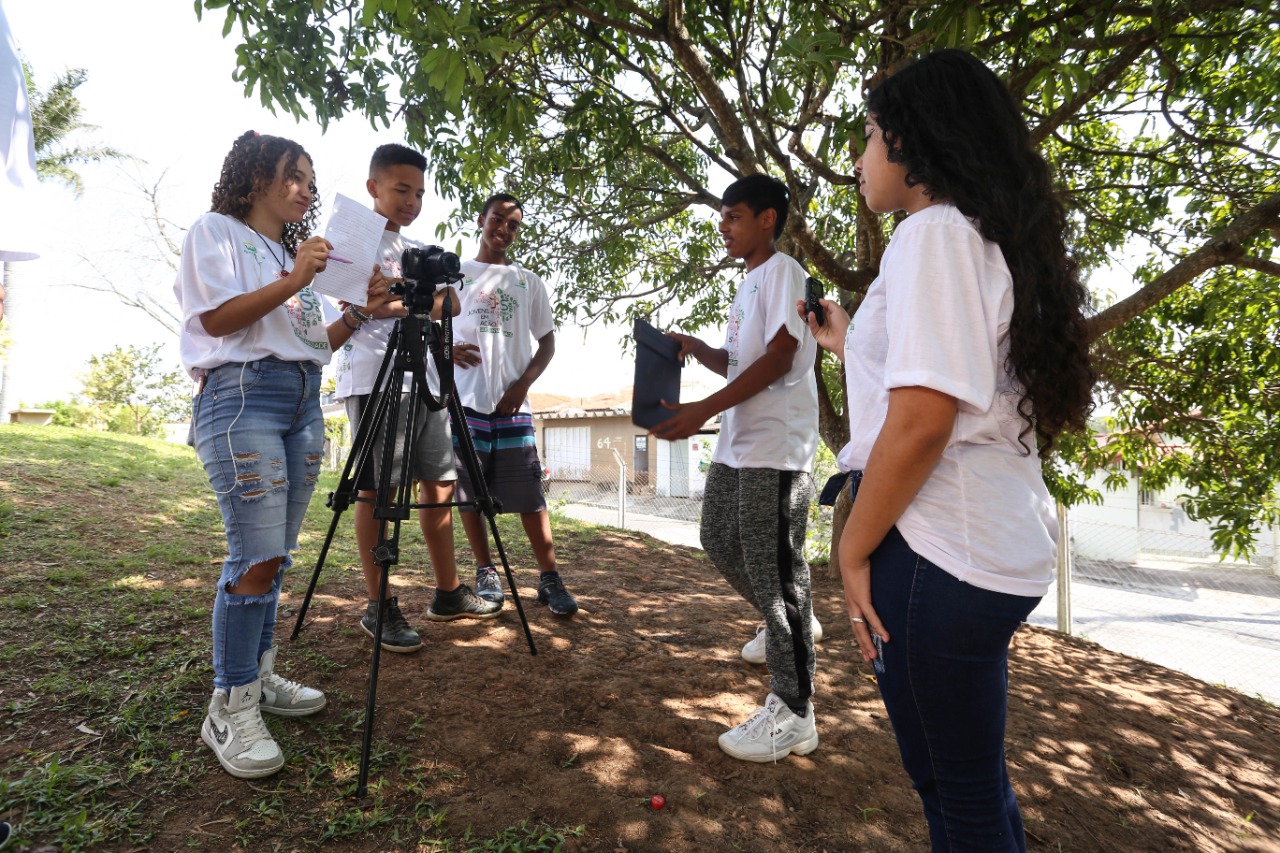 Grupo de alunos com camisetas brancas, da Associação Celebreiros, em volta de uma câmera, ao ar livre, com árvores em volta.