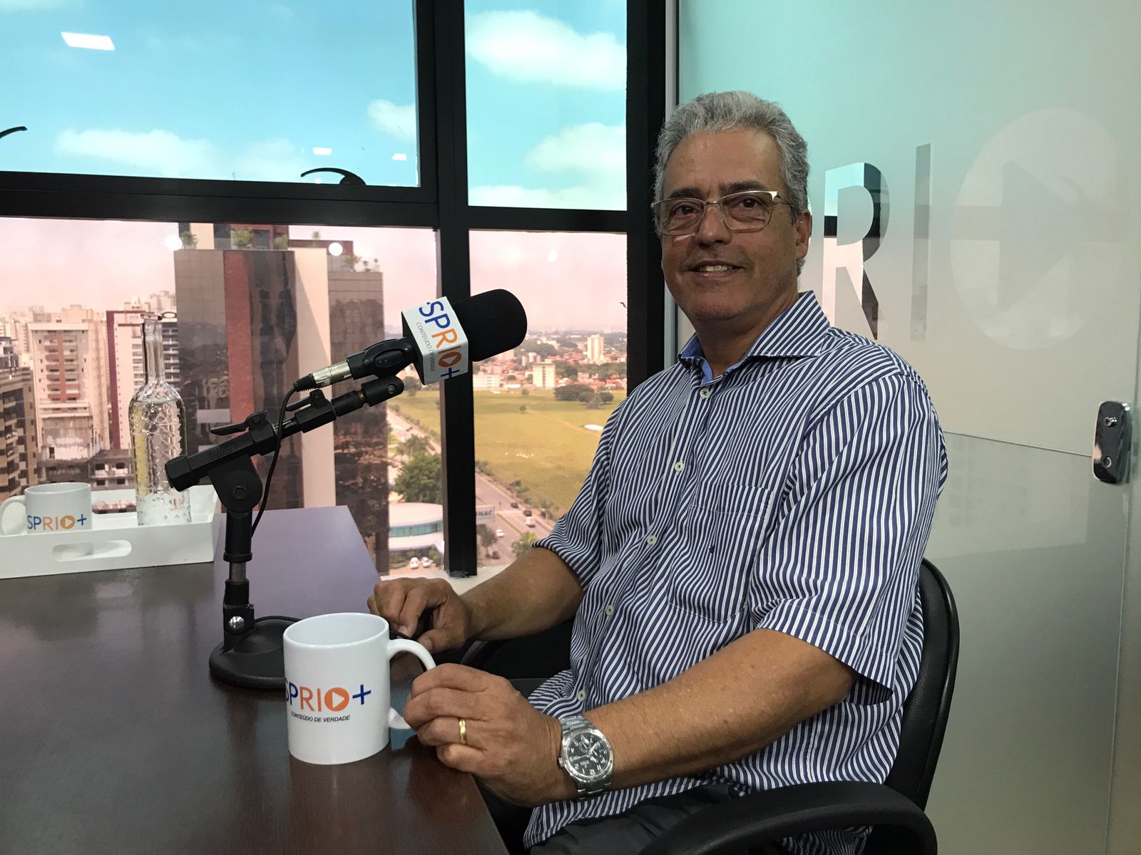 Retrato do Eduardo Falcão de Carvalho, da Estância SIlvânia, em Caçapava, sentado, falando ao microfone, com uma caneca nas mãos, janela ao fundo e vidro, falando sobre a Silvânia Week.