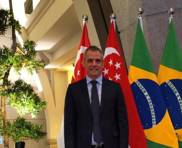 Retrato de Patrick Teyssonneyre em frente à bandeira do Brasil, com terno, gravata e sorrindo para a câmera.