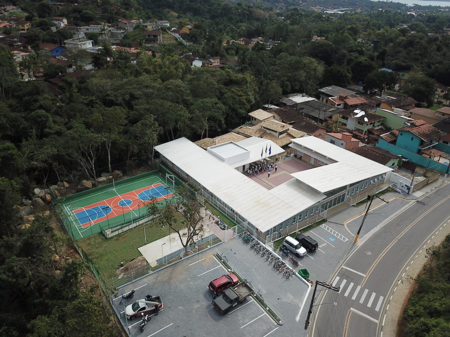 Vista aérea da Escola Cívico Municipal de Ilhabela, com o predio no meio, ruas em volta, carros estacionados e árvores ao fundo.