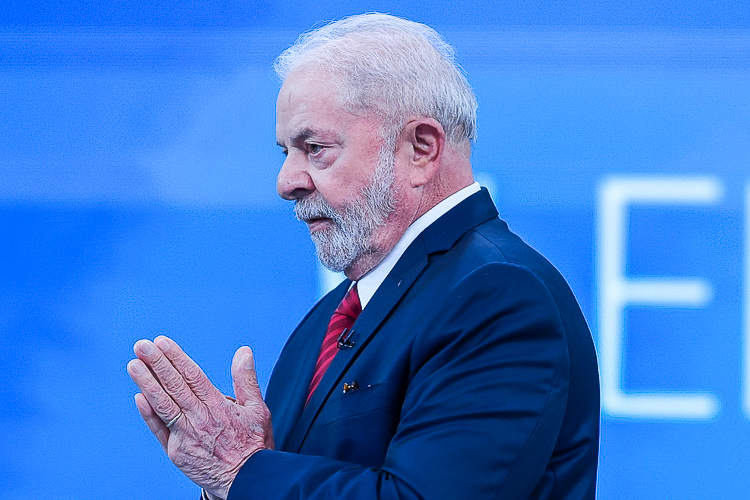 Lula tem alta do hospital Sírio-Libanês. Na foto, o petista está com as mãos juntas, tenro preto, gravata vermelha, olhando para a esquerda, com um fundo azul.