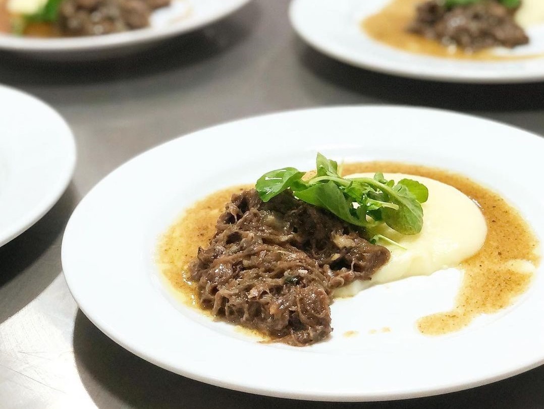Aligot da Escola de Gastronomia Chef Gourmet Taubaté, em um prato branco, com carne e queijo, em uma mesa.