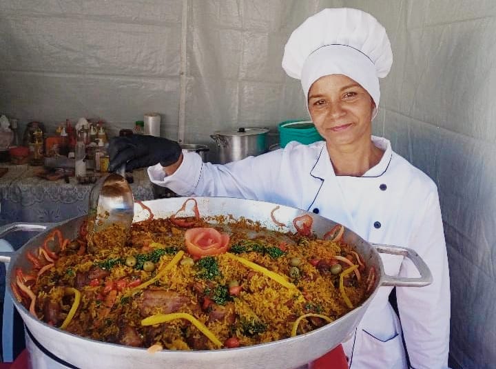 Chef da Arena Majestade SJC com roupa branca, chapéu de cozinheiro, com uma panela com comida, ingredientes ao fundo e parede cinza.