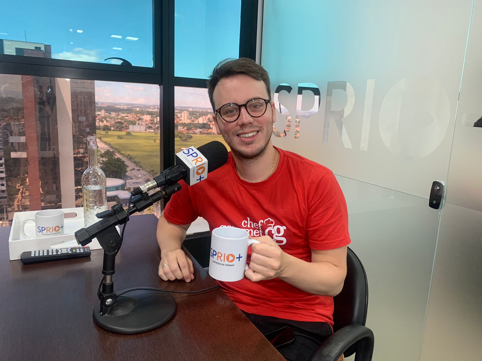 Rafael Ferro durante entrevista ao portal SP RIO+, segurando uma caneca, camiseta vermelha e vidro atrás.