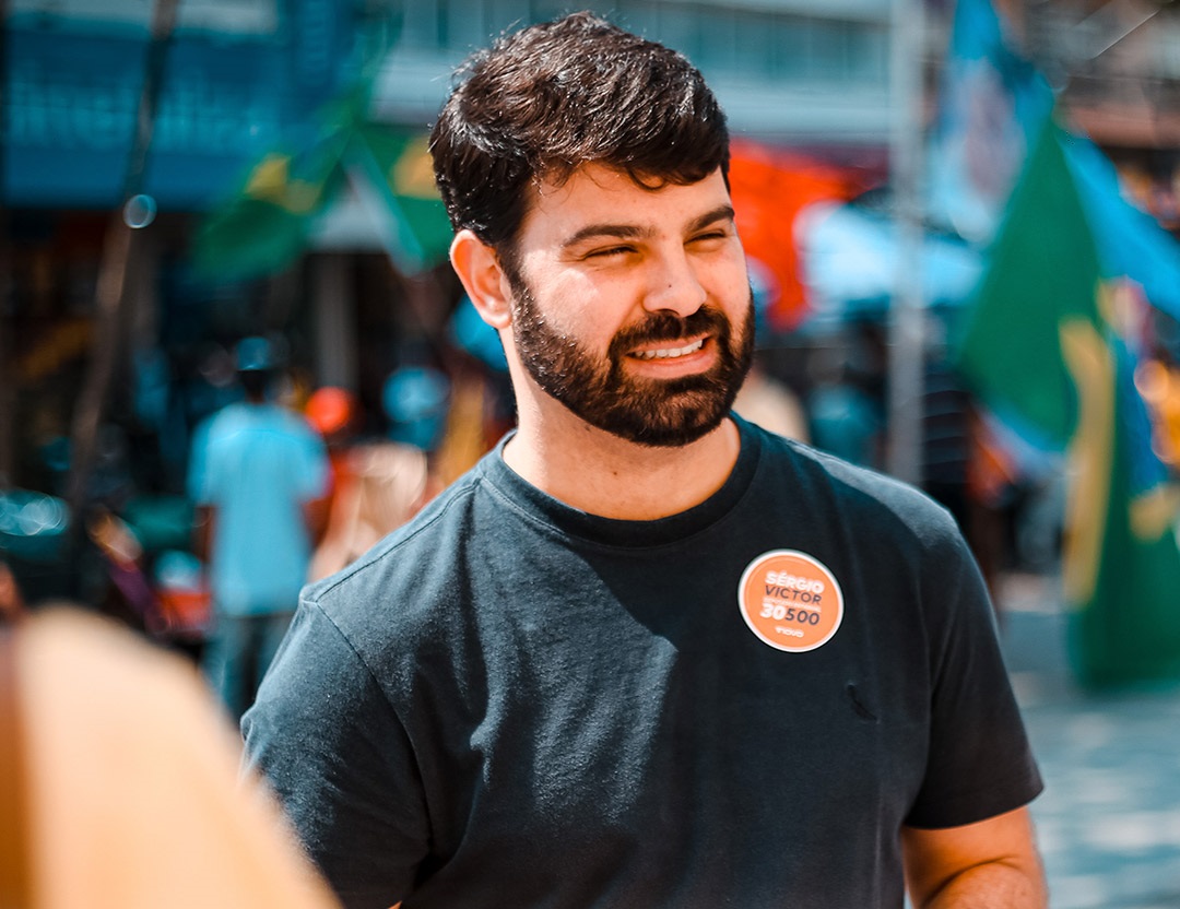 Sérgio Victor, deputado estadual, com uma camiseta preta, um selo do Partido Novo no peito, sorrindo, olhando para a direita, com o fundo desfocado de uma cidade.