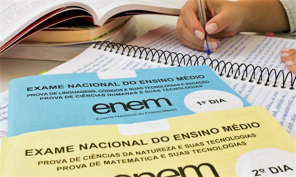 Cadernos do Enem, um azul e um amarelo, em cima de um caderno, com um mão escrevendo, simulando a matéria que mostra que o ENEM pode ser utilizado para ingressar em universidades portuguesas.