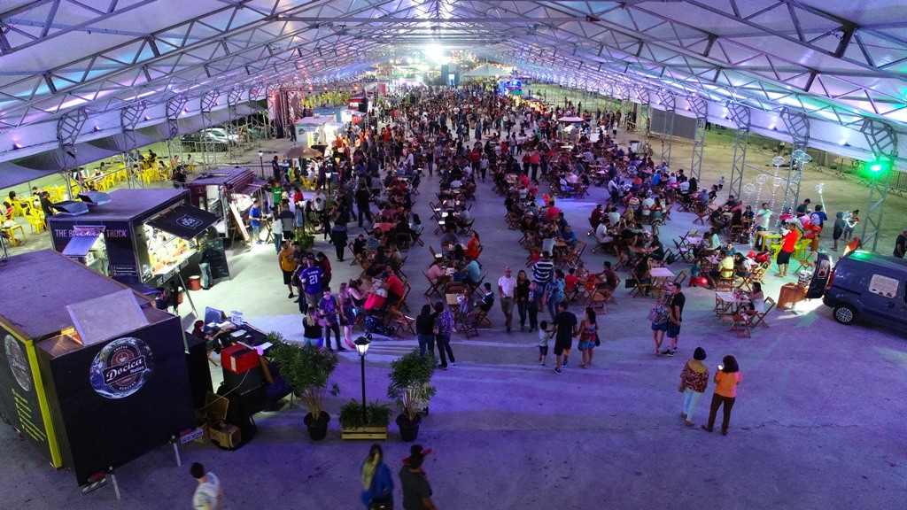 Pessoas ruenidas no Fetsival de Food Truck, em Caraguatatuba, sentadas, comendo, com trailers em volta e uma cobertura branca, de ferro.