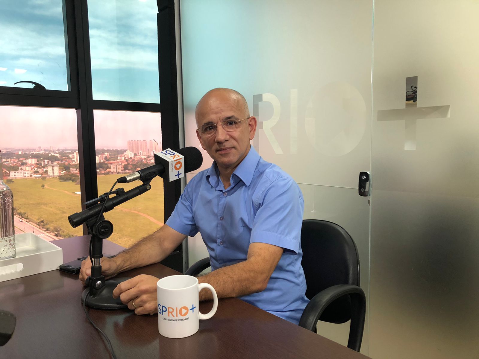 Robertinho da Padaria, presidente da Câmara de SJC, com uma camisa azul, falando ao microfone, uma caneca na mesa, a cidade de São José dos Campos ao fundo.