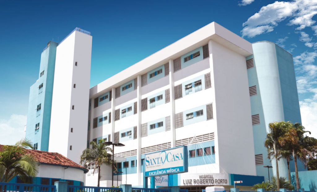 Fachada da Santa Casa SJC, com o prédio branco e azul, placa em baixo, janelas, céu azul e árvores em volta.