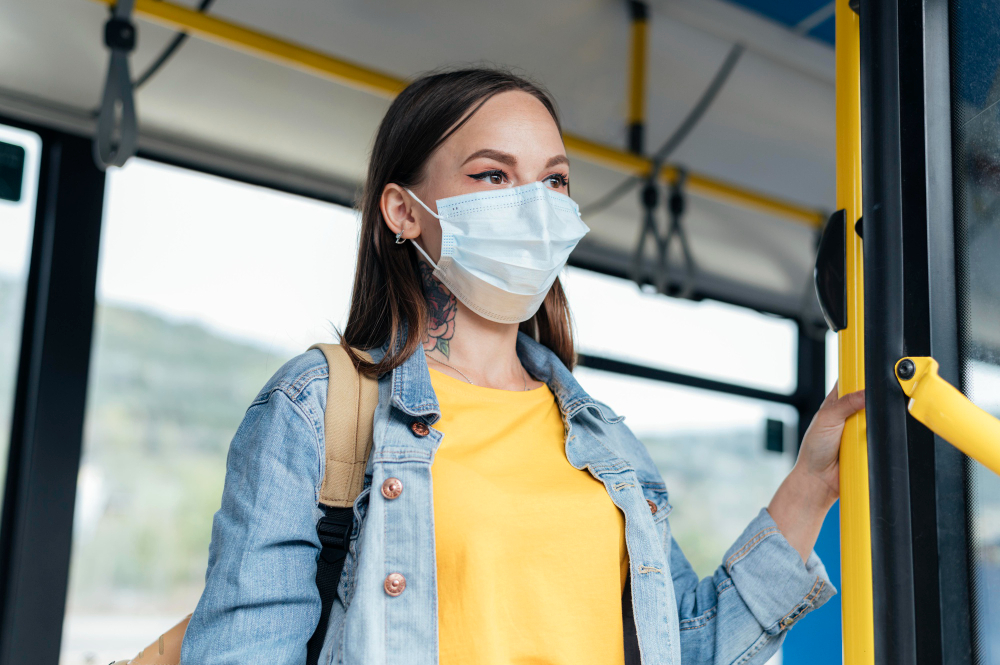 Mulher com uma máscara branca em um transporte público, segurando em uma barra, com uma jaqueta jeans, camisa amarela, olhando para fora. Foto ilustra a matéria das máscaras em Caçapava.