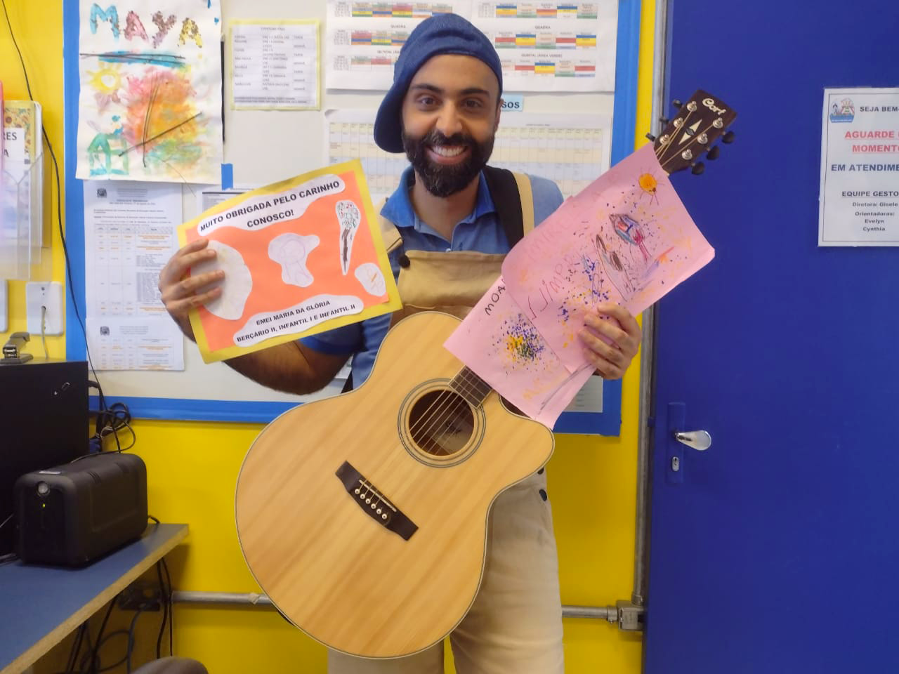 Tiago Vilela, música de SJC, segurando um violão, com cartazes nas mãos, em uma escola da cidade, com parede amarela e azul.