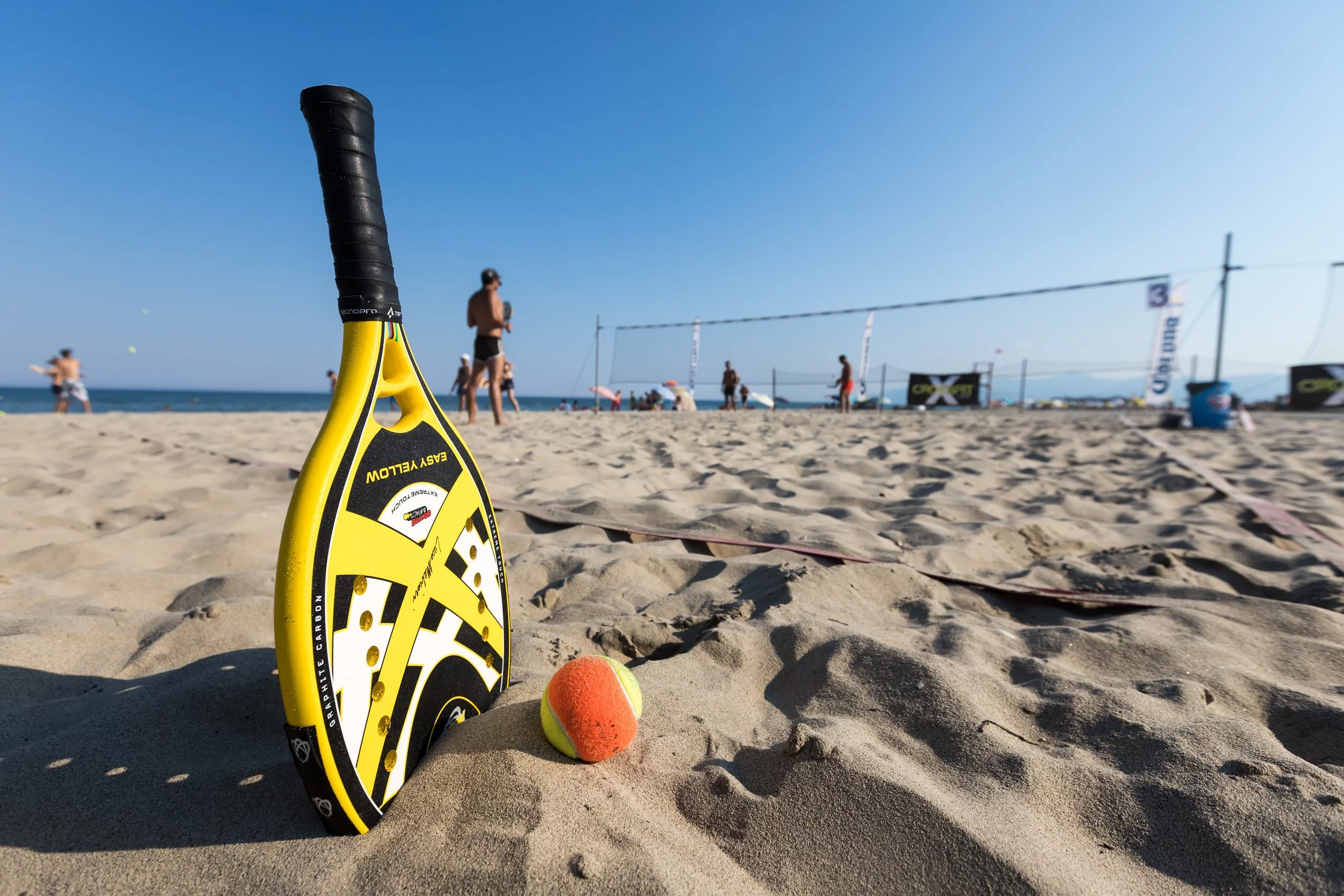 Foto mostra uma raquete na areia, ao lado de uma bola, simulando a matéria do torneio de beach tennis em São Sebastião, o 1° São Sebá Open, com uma rede ao fundo, céu azul e parte do mar.