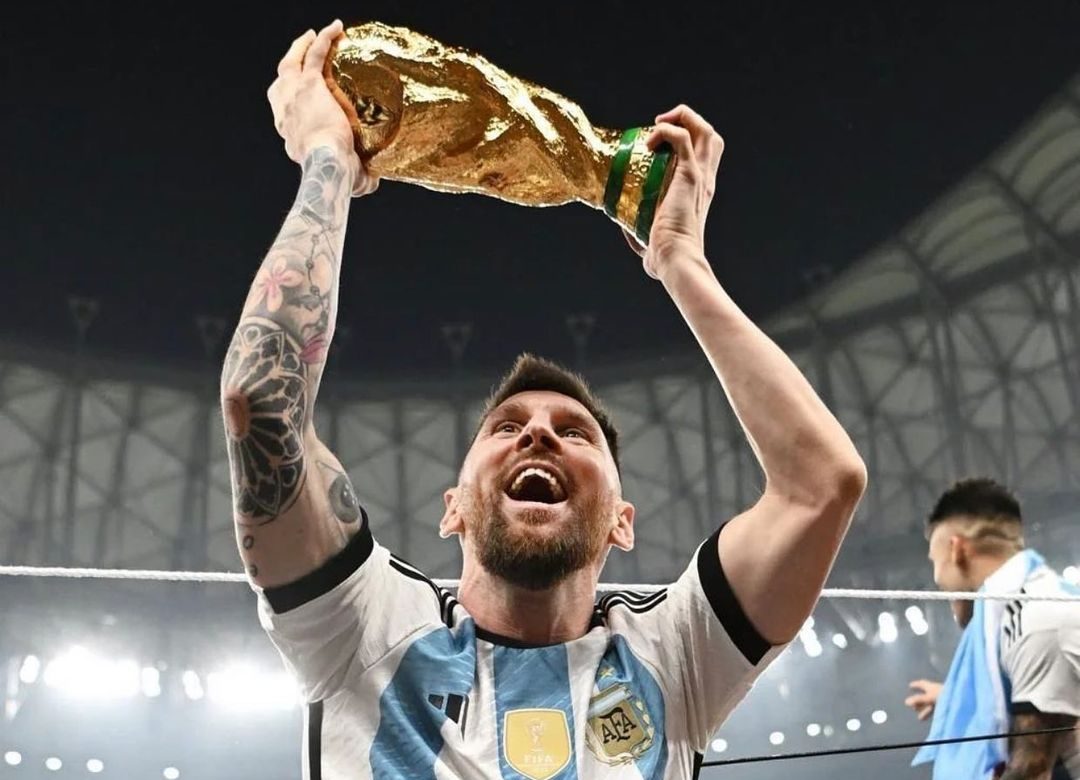 messi sorri segurando a taça da copa do mundo no alto com as duas mãos